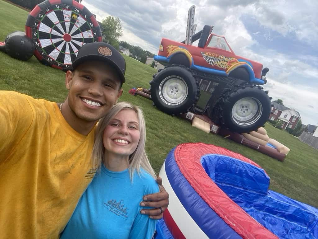 Party Bounce House Rentals of Knoxville team smiling at a Knoxville backyard party with dart wall, monster truck inflatable, and water slide setup in the background